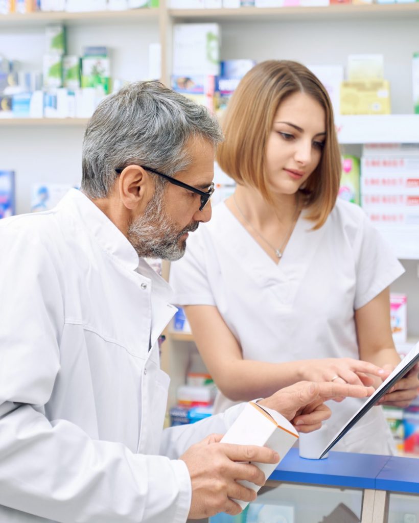 Mature bearded man in eyeglasses talking, consulting with female pharmacist in drugstore. Woman and man in white lab coats holding black folder, checking and providing informaion about medicines.